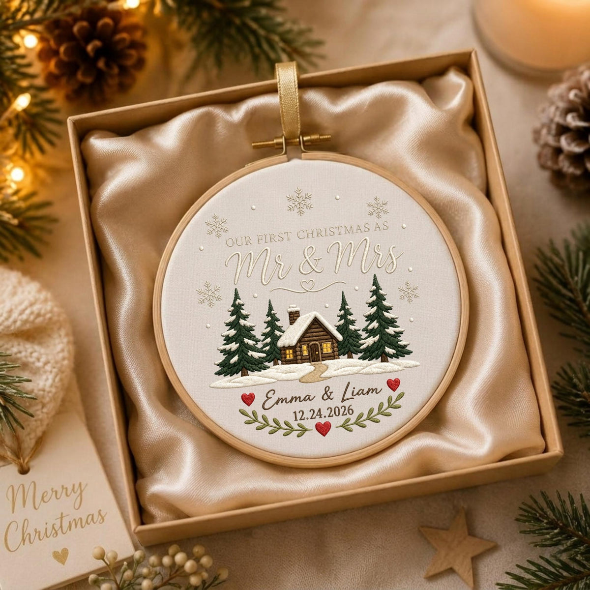 May include: A round embroidery hoop ornament featuring a white fabric center with a stitched winter cabin scene, pine trees, and snowflakes. The text reads "OUR FIRST CHRISTMAS AS Mr &amp; Mrs" followed by "Emma &amp; Liam" and "12.24.2026".