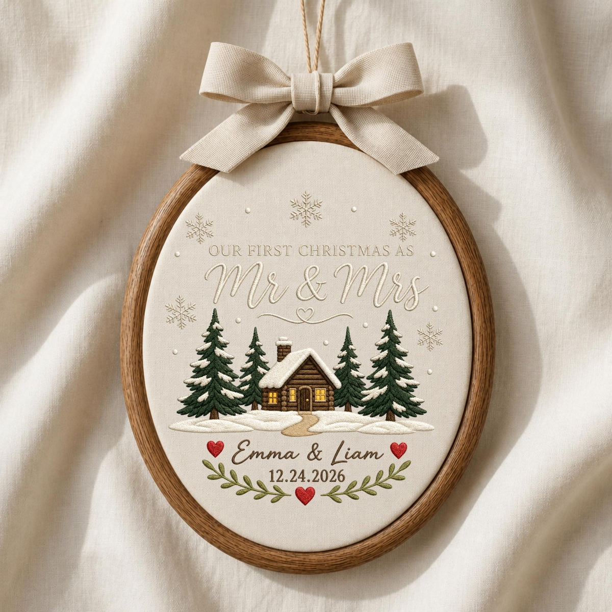 May include: An oval wooden embroidery hoop frame containing fabric with embroidered text "OUR FIRST CHRISTMAS AS Mr &amp; Mrs", "Emma &amp; Liam", and "12.24.2026". The design features a cabin, pine trees, snowflakes, and red hearts, topped with a beige ribbon.