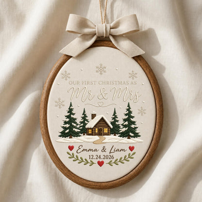 May include: An oval wooden embroidery hoop frame containing fabric with embroidered text "OUR FIRST CHRISTMAS AS Mr &amp; Mrs", "Emma &amp; Liam", and "12.24.2026". The design features a cabin, pine trees, snowflakes, and red hearts, topped with a beige ribbon.