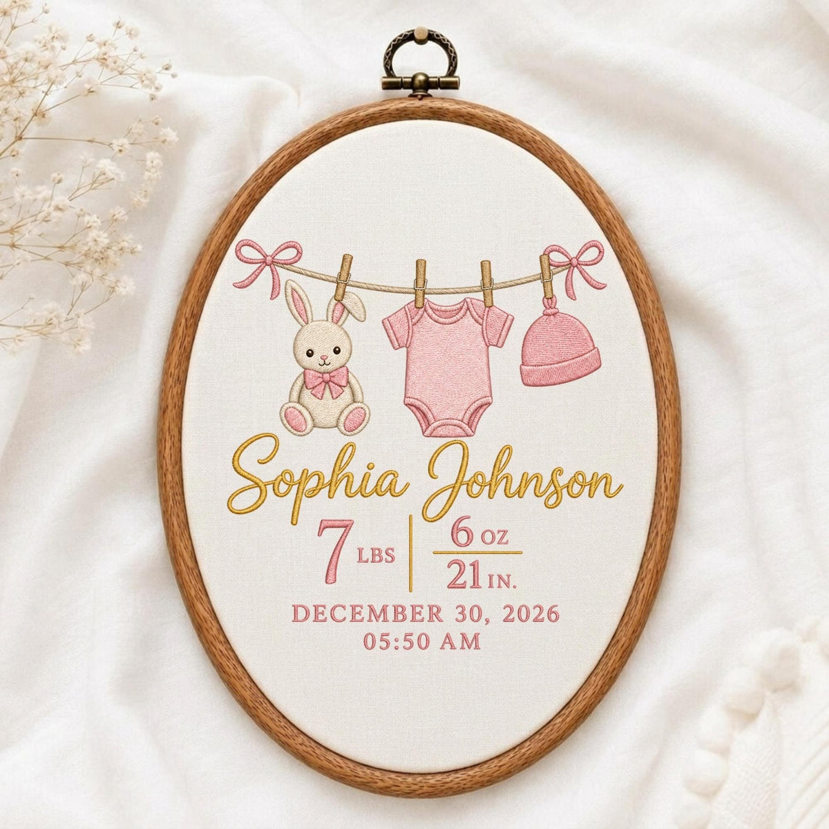 May include: An oval embroidery hoop containing fabric with a stitched design of a bunny, a onesie, and a hat hanging on a line. The text reads Sophia Johnson, 7 lbs 6 oz, 21 in., December 30, 2026, and 05:50 AM in pink and gold thread.