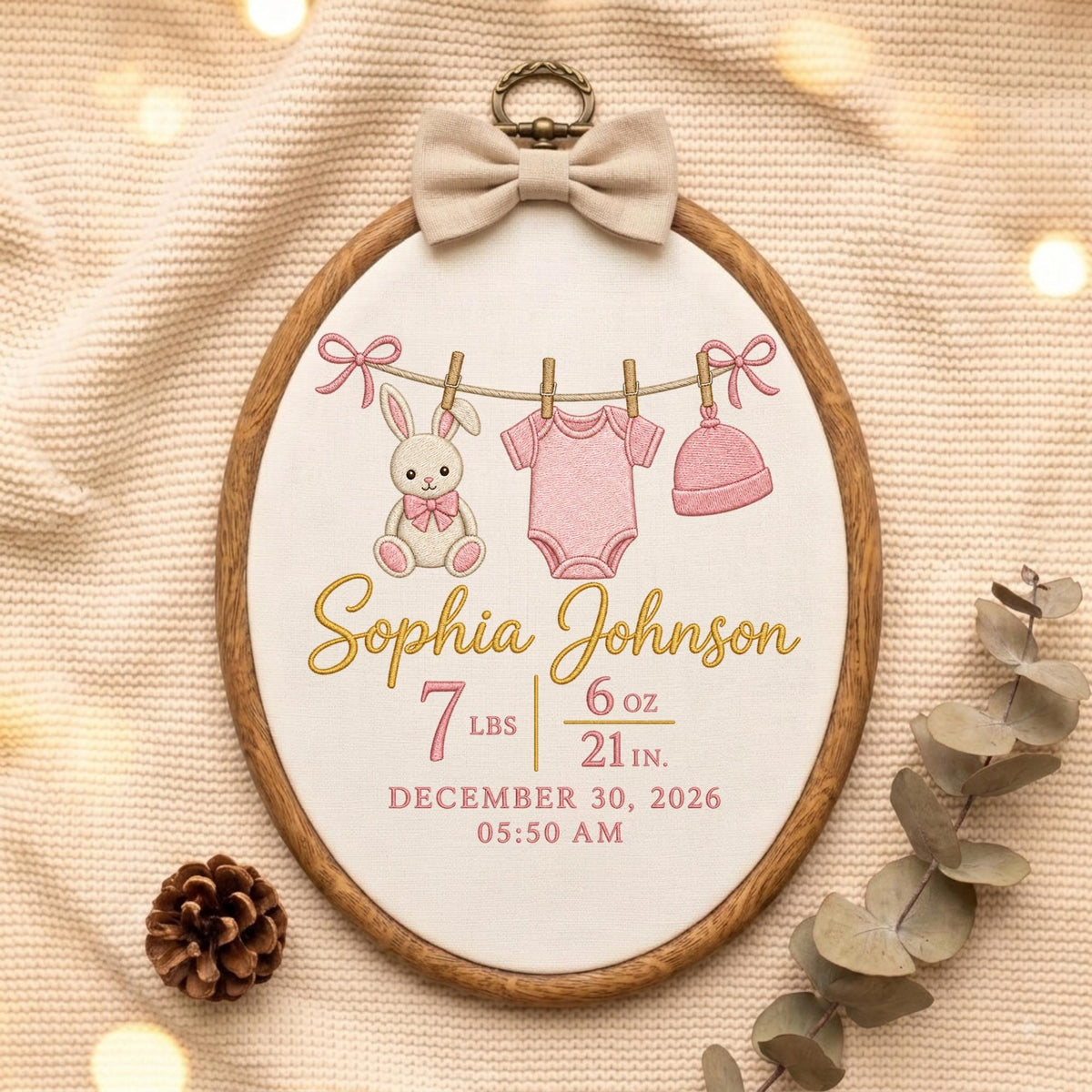 May include: An oval wooden embroidery hoop featuring a cream fabric background with a beige bow at the top. The embroidery depicts a bunny, a pink baby bodysuit, and a pink hat hanging on a line. Text includes Sophia Johnson, 7 lbs 6 oz, 21 in, December 30, 2026, and 05:50 AM.