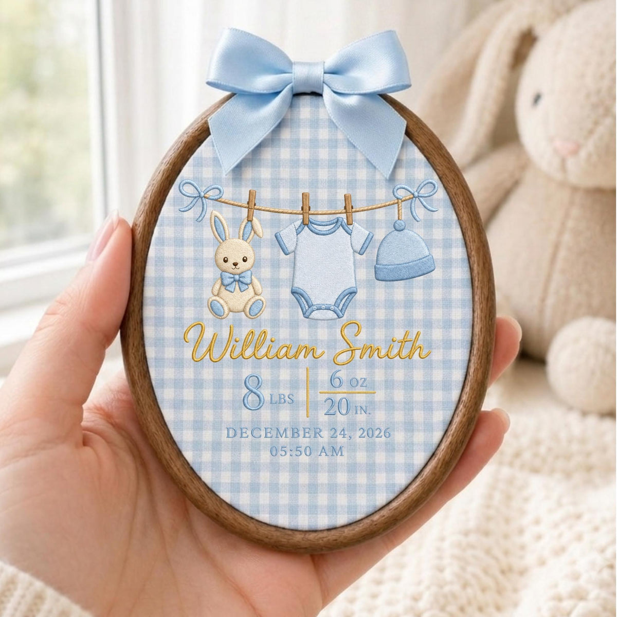 May include: An oval wooden embroidery hoop featuring a light blue gingham fabric background. It displays an embroidered clothesline with a bunny, a onesie, and a hat. Text reads: William Smith, 8 lbs 6 oz, 20 in., December 24, 2026, 05:50 AM. A light blue satin bow is attached at the top.