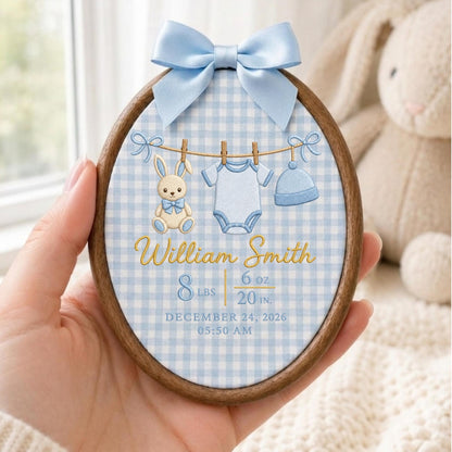 May include: An oval wooden embroidery hoop featuring a light blue gingham fabric background. It displays an embroidered clothesline with a bunny, a onesie, and a hat. Text reads: William Smith, 8 lbs 6 oz, 20 in., December 24, 2026, 05:50 AM. A light blue satin bow is attached at the top.