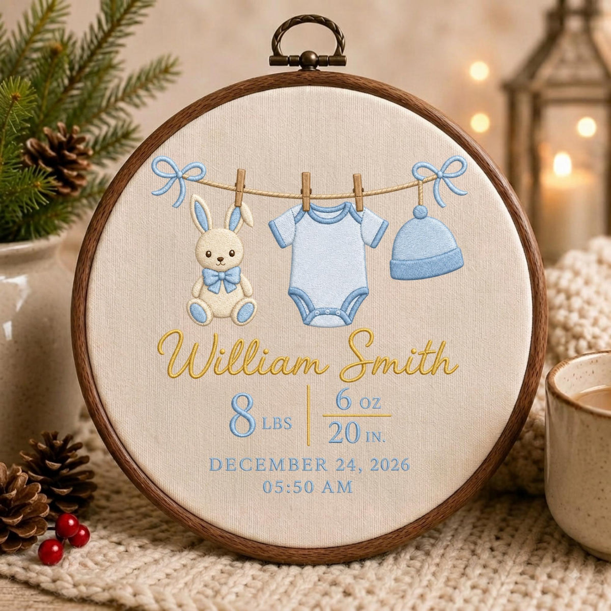 May include: A circular wooden embroidery hoop containing cream fabric with embroidered baby items: a beige rabbit, a light blue onesie, and a light blue hat hanging on a line. Text reads: William Smith, 8 lbs 6 oz, 20 in, December 24, 2026, 05:50 AM.