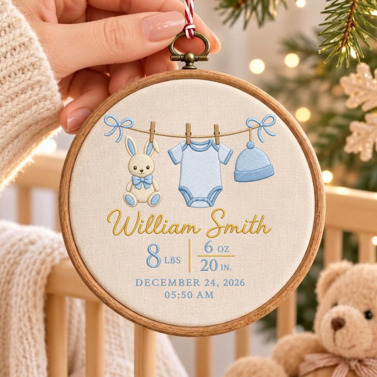 May include: A circular wooden embroidery hoop containing cream fabric with a stitched design of a bunny, a blue onesie, and a blue hat hanging on a line. The text reads William Smith, 8 lbs 6 oz, 20 in, December 24, 2026, 05:50 AM.