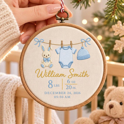 May include: A circular wooden embroidery hoop containing cream fabric with a stitched design of a bunny, a blue onesie, and a blue hat hanging on a line. The text reads William Smith, 8 lbs 6 oz, 20 in, December 24, 2026, 05:50 AM.