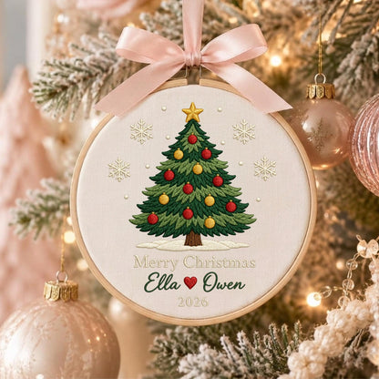 May include: A round wooden embroidery hoop ornament featuring a cream fabric background with a stitched green Christmas tree decorated with red and yellow ornaments, topped with a gold star, surrounded by snowflakes, and the text Merry Christmas Ella and Owen 2026 with a red heart, finished with a light pink satin ribbon bow.