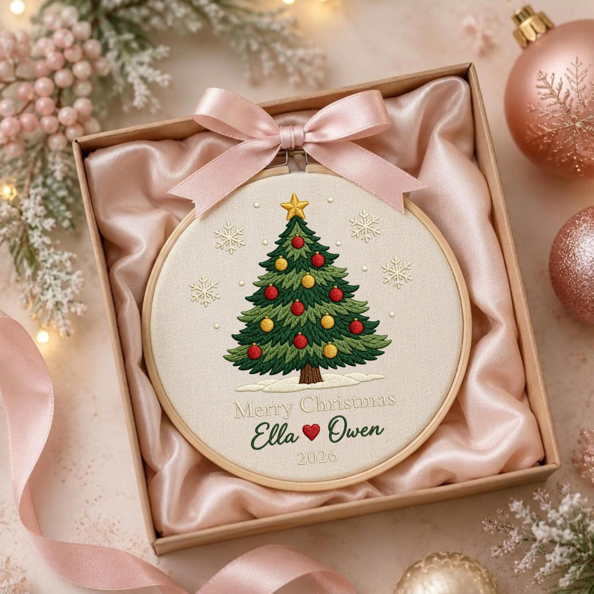 May include: A round embroidery hoop containing a fabric panel with a stitched green Christmas tree, red and yellow ornaments, and a gold star. The text reads Merry Christmas, Ella, a red heart, Owen, and 2026. A pink satin ribbon is tied at the top.
