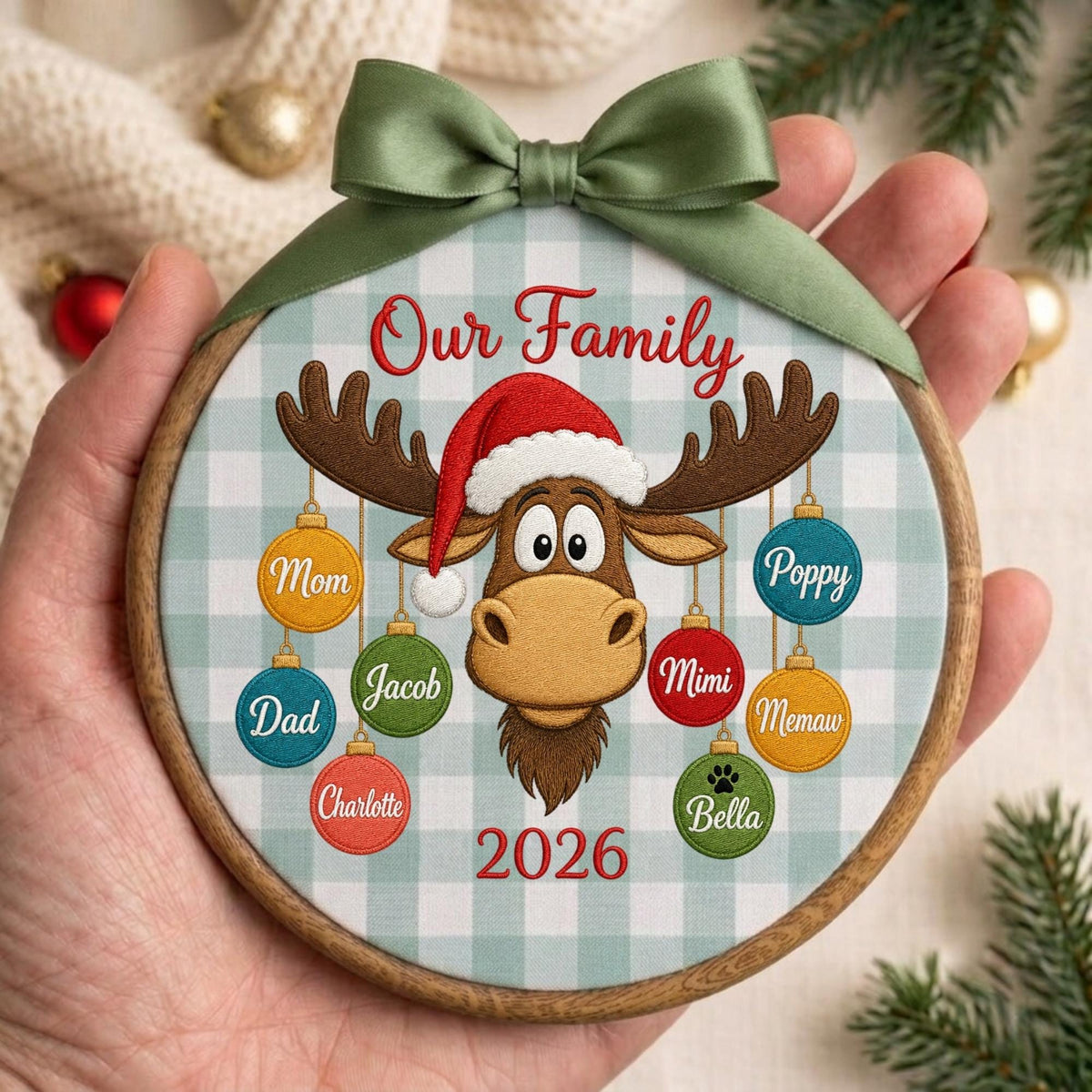 May include: A round wooden embroidery hoop ornament featuring a light blue and white buffalo check fabric. It displays an embroidered moose wearing a red Santa hat, surrounded by colorful hanging ornaments labeled with names and the year 2026.