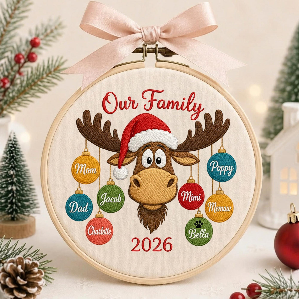 May include: A circular embroidery hoop featuring a light fabric background with a brown moose wearing a red Santa hat. The text Our Family and 2026 are embroidered in red. Eight colorful ornaments hang from the moose, each labeled with names.