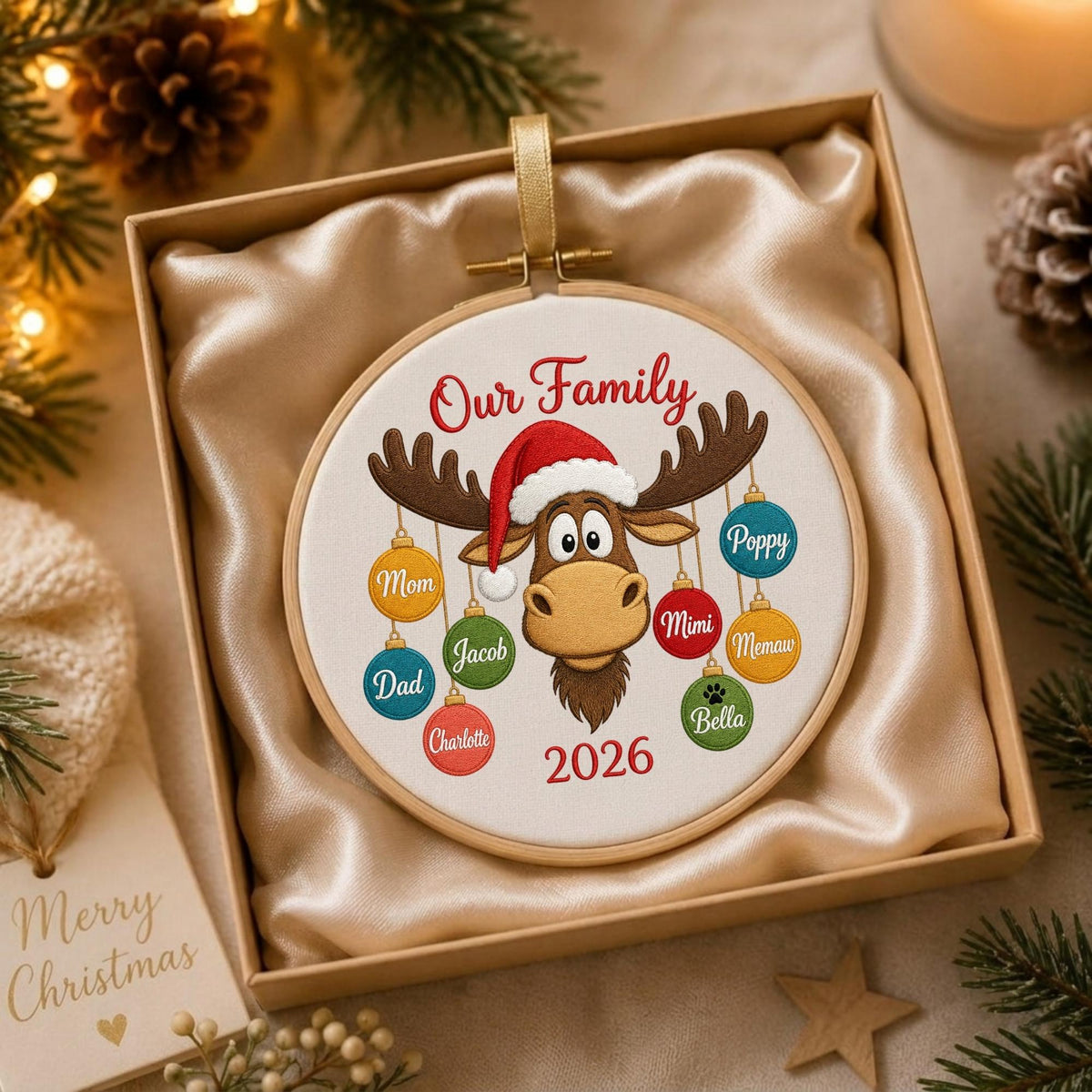 May include: A round wooden embroidery hoop ornament featuring a cartoon moose wearing a red Santa hat. The text reads Our Family and 2026. Hanging from the moose antlers are colorful Christmas ornaments labeled Mom, Dad, Charlotte, Jacob, Mimi, Memaw, Poppy, and Bella with a paw print.