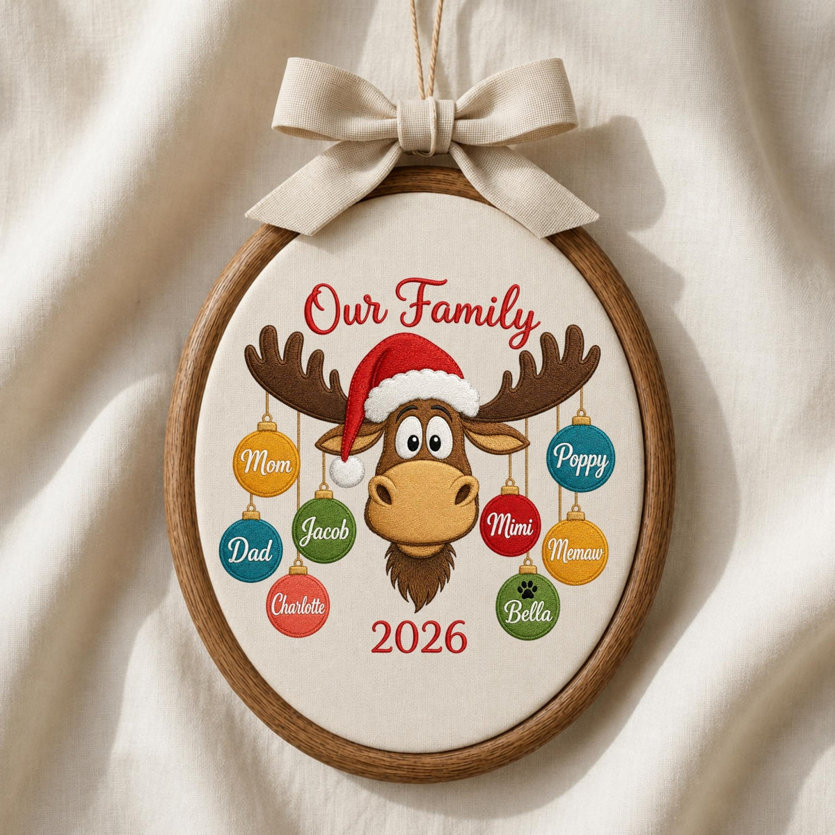May include: An oval wooden embroidery hoop ornament featuring a cream fabric center with a brown moose wearing a red Santa hat. The text reads Our Family and 2026. Eight colorful hanging ornaments are labeled with names: Mom, Dad, Charlotte, Jacob, Poppy, Mimi, Memaw, and Bella. A beige ribbon bow is attached at the top.