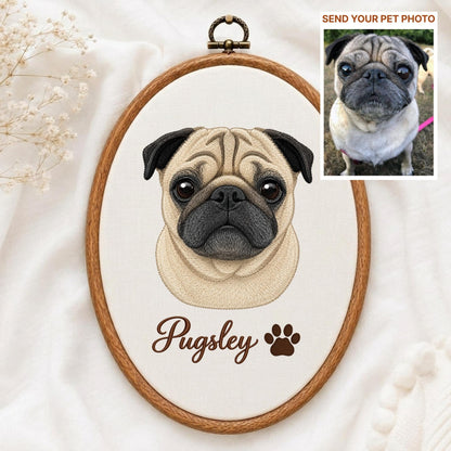 May include: An oval wooden embroidery hoop containing a stitched portrait of a fawn pug with a black muzzle and large dark eyes. Below the dog, the name Pugsley is embroidered in brown script next to a brown paw print icon on white fabric.