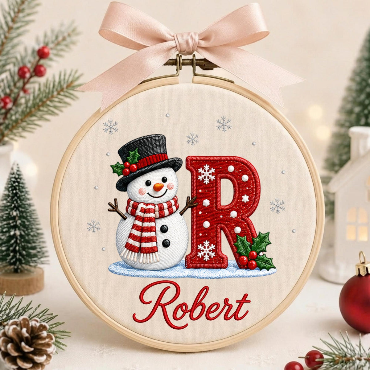 May include: A circular wooden embroidery hoop containing fabric with a stitched snowman wearing a black top hat and red and white striped scarf next to a large red letter R decorated with white snowflakes and holly, with the name Robert stitched below.