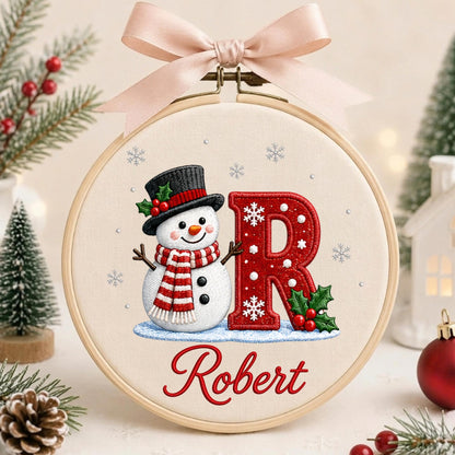 May include: A circular wooden embroidery hoop containing fabric with a stitched snowman wearing a black top hat and red and white striped scarf next to a large red letter R decorated with white snowflakes and holly, with the name Robert stitched below.