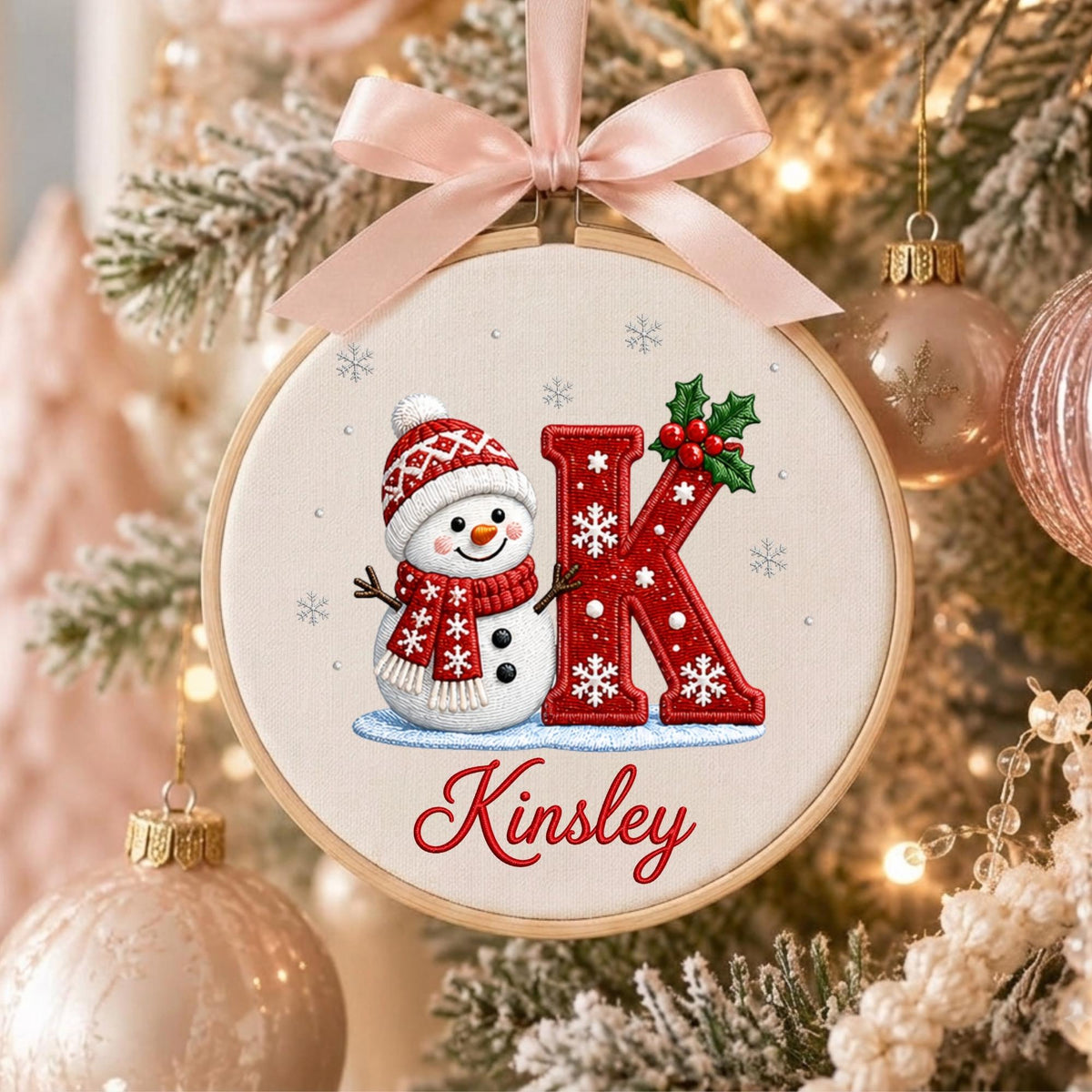 May include: A round wooden embroidery hoop ornament featuring a white fabric background with a snowman wearing a red patterned hat and scarf, a large red letter K decorated with white snowflakes and holly, and the name Kinsley in red script.