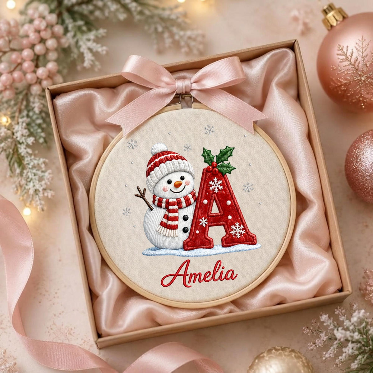 May include: A circular embroidery hoop featuring a white snowman wearing a red and white striped hat and scarf, next to a large red letter A decorated with white snowflakes and green holly leaves, with the name Amelia embroidered in red script below.