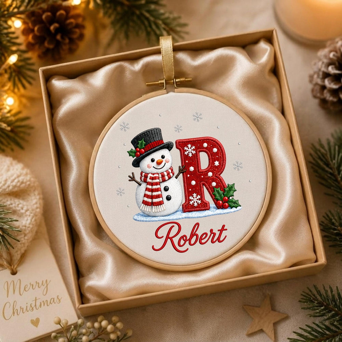 May include: A circular embroidery hoop ornament featuring a white fabric background with a snowman wearing a black hat and red striped scarf next to a large red letter R decorated with white snowflakes and green holly leaves, with the name Robert.