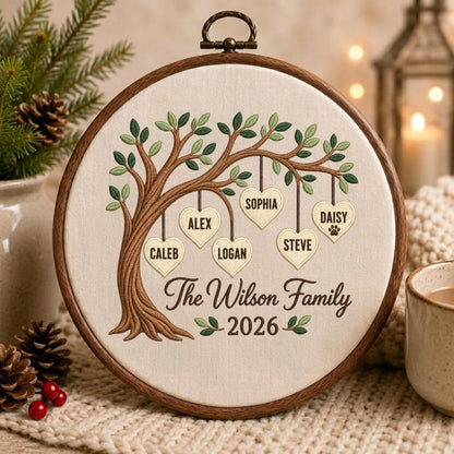 May include: A circular wooden embroidery hoop containing a cream-colored fabric with a brown embroidered tree. Hanging from the branches are six cream-colored hearts with the names Caleb, Alex, Logan, Sophia, Steve, and Daisy. Text reads The Wilson Family 2026.
