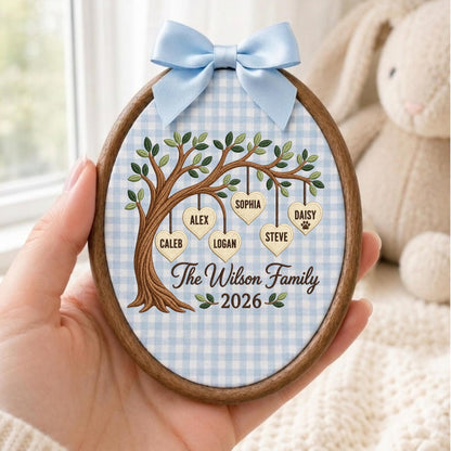 May include: An oval wooden embroidery hoop frame containing fabric with a light blue gingham pattern. An embroidered tree features hanging heart-shaped tags with the names Caleb, Alex, Sophia, Logan, Steve, and Daisy. Text reads The Wilson Family 2026.