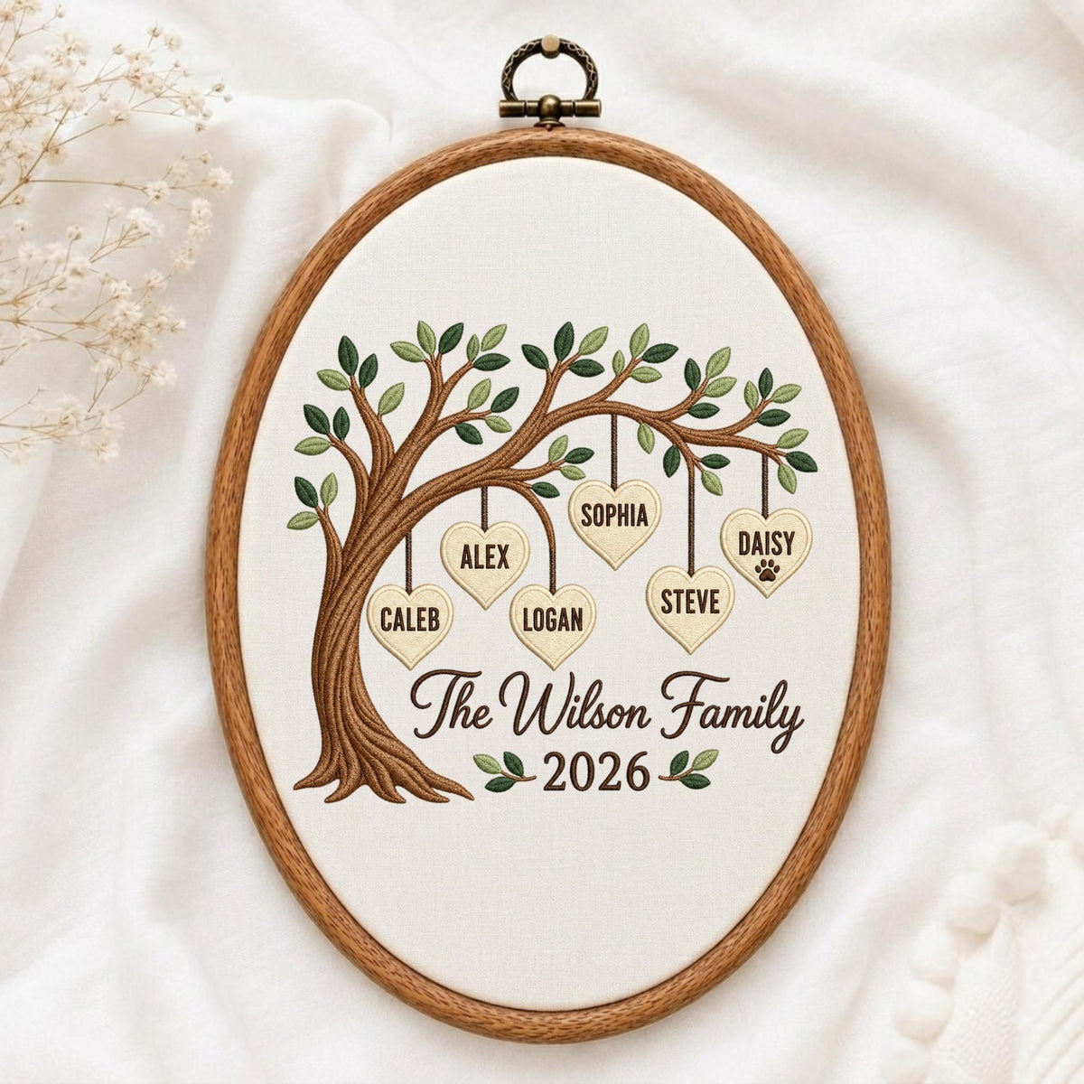 May include: An oval embroidery hoop containing a fabric panel with a stitched brown tree. Hanging from the branches are five cream-colored heart shapes labeled Alex, Sophia, Daisy, Steve, and Logan. The text reads The Wilson Family 2026.