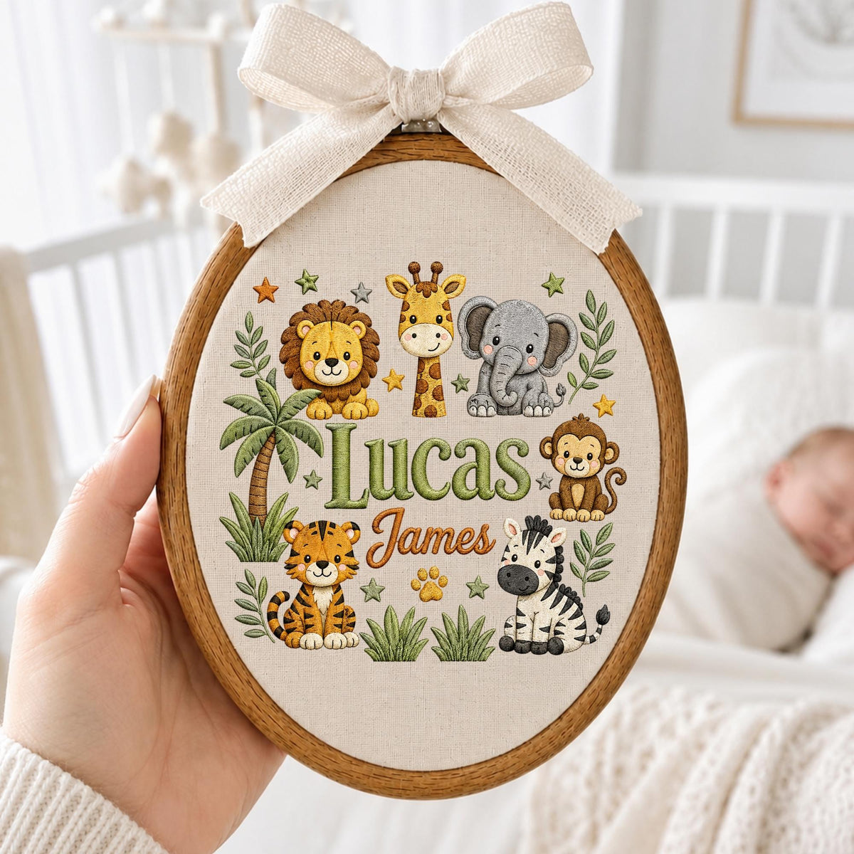 May include: An oval wooden embroidery hoop featuring a cream fabric background with a beige ribbon bow at the top. The design includes embroidered illustrations of a lion, giraffe, elephant, monkey, tiger, and zebra, with the text Lucas James.