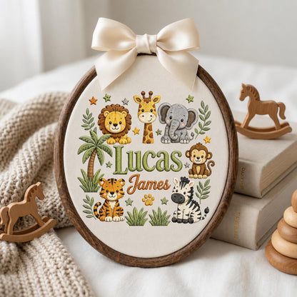 May include: An oval embroidery hoop with a dark wood frame and a cream fabric center. The fabric features embroidered jungle animals including a lion, giraffe, elephant, monkey, tiger, and zebra, with the names Lucas James and a palm tree.