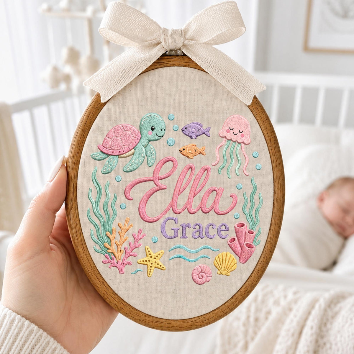 May include: An oval wooden embroidery hoop containing fabric with a stitched underwater scene featuring a pink and green sea turtle, a pink jellyfish, small fish, coral, a starfish, and seashells. The name Ella Grace is embroidered in pink and purple.