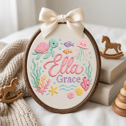 May include: An oval embroidery hoop featuring a cream fabric background with a stitched sea life design, including a green turtle, pink jellyfish, starfish, and coral. The name Ella Grace is embroidered in pink and purple script with a cream bow.