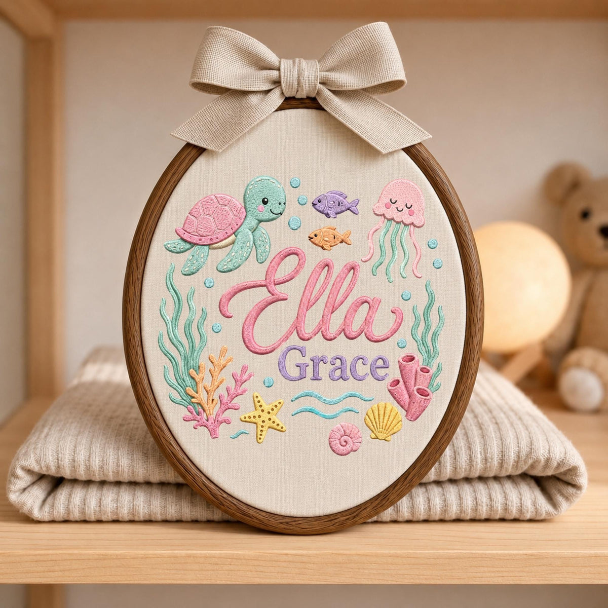 May include: An oval embroidery hoop featuring a cream fabric background with the name Ella Grace stitched in pink and purple. The design includes a green sea turtle, a pink jellyfish, coral, a yellow starfish, and small fish in various colors.