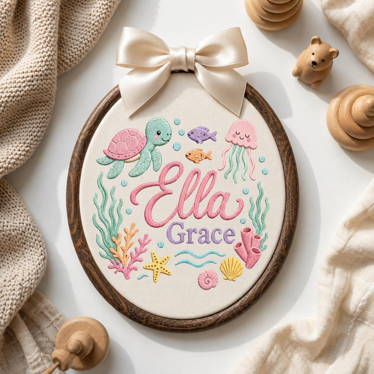 May include: An oval embroidery hoop featuring a cream fabric background with a colorful ocean-themed design. The embroidery includes a pink turtle, a jellyfish, fish, coral, and a starfish. The name Ella Grace is stitched in pink and purple script.