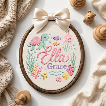 May include: An oval embroidery hoop featuring a cream fabric background with a colorful ocean-themed design. The embroidery includes a pink turtle, a jellyfish, fish, coral, and a starfish. The name Ella Grace is stitched in pink and purple script.