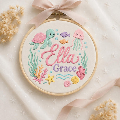 May include: An embroidery hoop featuring a light-colored fabric with a stitched underwater scene. The design includes a pink and teal turtle, a pink jellyfish, fish, coral, a starfish, and shells. The name Ella Grace is embroidered in pink and purple.