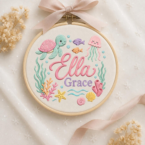 May include: An embroidery hoop featuring a light-colored fabric with a stitched underwater scene. The design includes a pink and teal turtle, a pink jellyfish, fish, coral, a starfish, and shells. The name Ella Grace is embroidered in pink and purple.