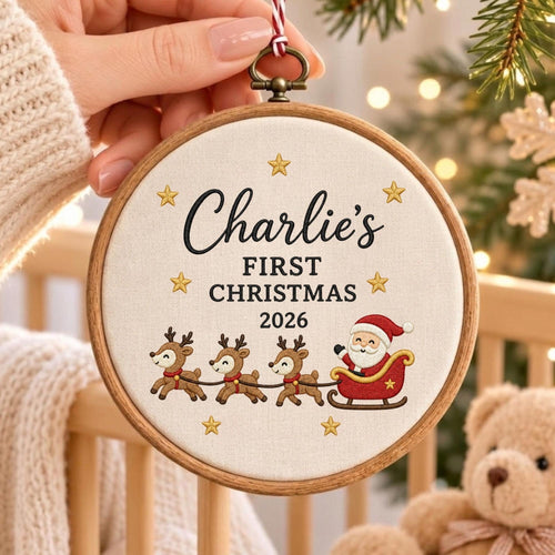 May include: A round wooden embroidery hoop containing off-white fabric with embroidered text reading Charlie's FIRST CHRISTMAS 2026. The design features six small gold stars and a red sleigh pulled by three brown reindeer with Santa Claus.