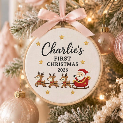 May include: A round wooden embroidery hoop ornament featuring white fabric with the embroidered text Charlie's FIRST CHRISTMAS 2026, gold stars, three reindeer, and Santa in a red sleigh, topped with a light pink satin ribbon bow.
