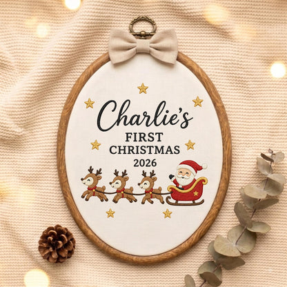 May include: An oval wooden embroidery hoop containing white fabric with the embroidered text Charlie's FIRST CHRISTMAS 2026. The design features a red sleigh with Santa Claus, three brown reindeer, and gold stars. A beige fabric bow sits at the top.
