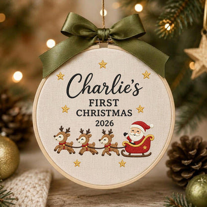 May include: A round embroidery hoop ornament featuring a cream-colored fabric background with the embroidered text Charlie's FIRST CHRISTMAS 2026, surrounded by gold stars, three reindeer, and Santa in a red sleigh, topped with a dark olive green bow.