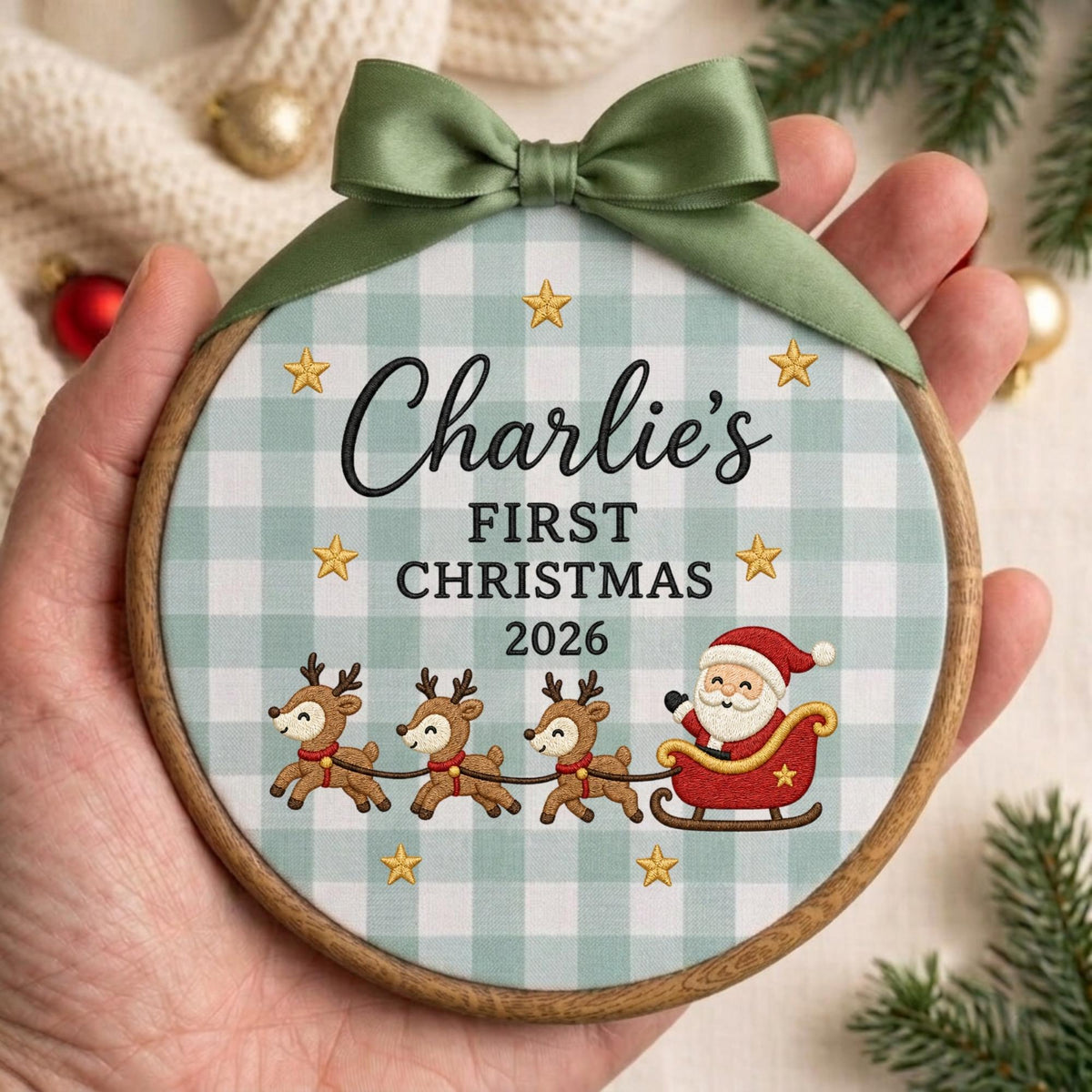 May include: A round wooden embroidery hoop ornament featuring a light green and white gingham fabric background. The text reads Charlie's FIRST CHRISTMAS 2026. It includes a graphic of Santa Claus in a sleigh pulled by three reindeer and gold stars.