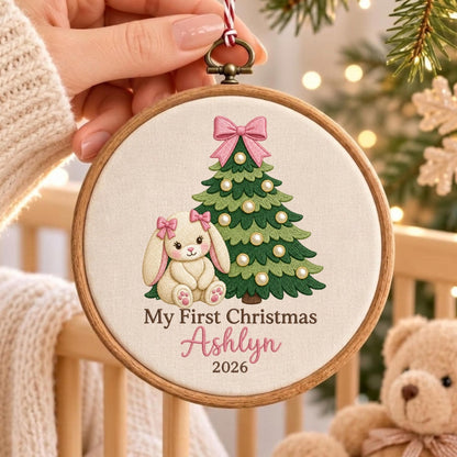 May include: A round wooden embroidery hoop containing cream-colored fabric with an embroidered green Christmas tree, a cream-colored bunny with pink bows, and the text My First Christmas Ashlyn 2026 in brown and pink script lettering.