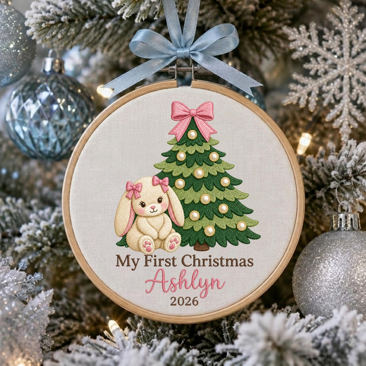 May include: A round wooden embroidery hoop ornament featuring a cream-colored fabric center. The design includes an embroidered green Christmas tree with white pearl-like accents, a beige bunny with pink bows, and the text My First Christmas Ashlyn 2026.