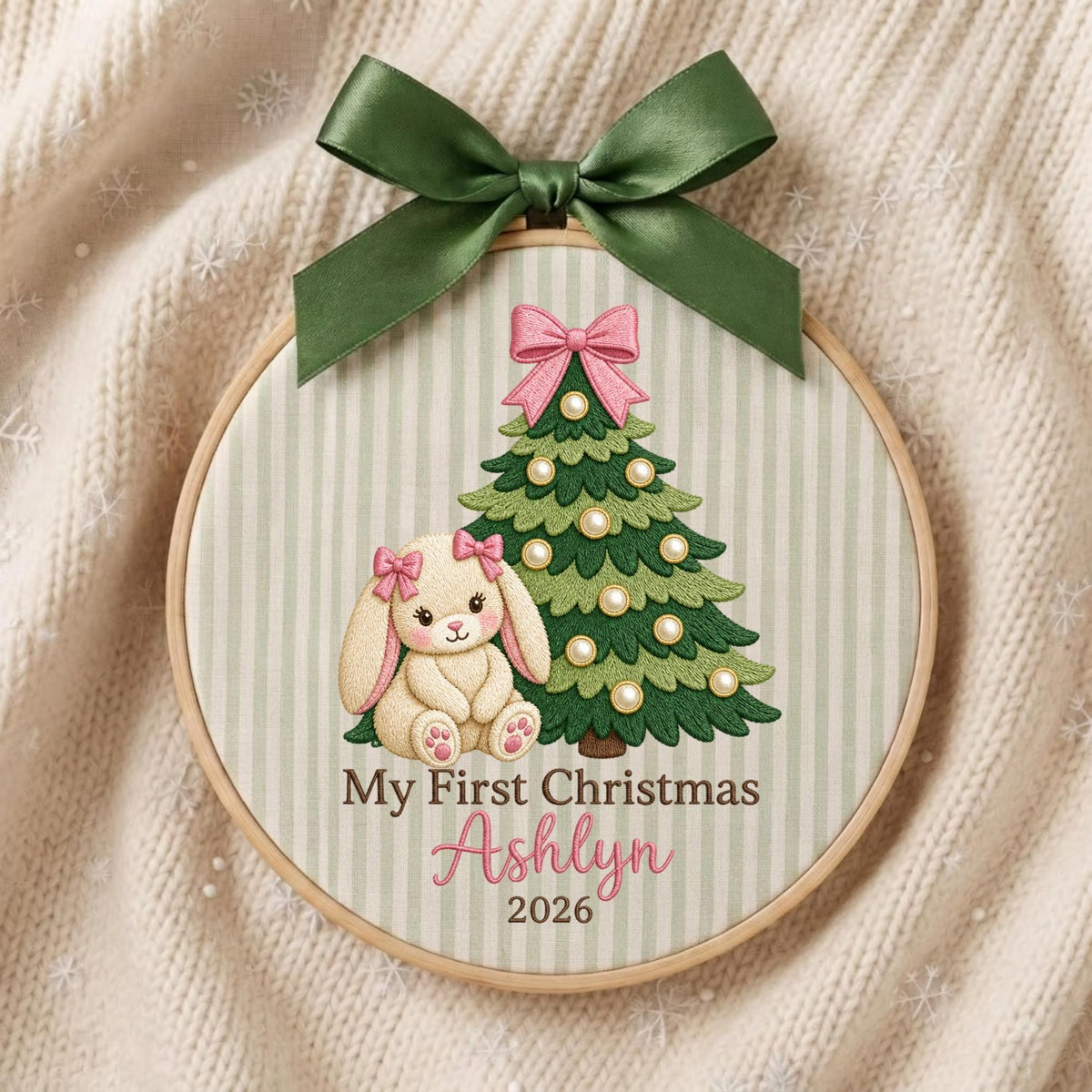 May include: A round wooden embroidery hoop containing fabric with a light green striped pattern. It features an embroidered green Christmas tree with white ornaments, a cream-colored bunny with pink bows, and the text My First Christmas Ashlyn 2026.
