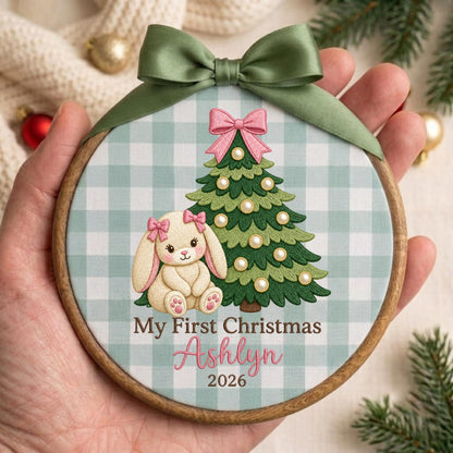 May include: A round wooden embroidery hoop ornament featuring a light green and white gingham fabric background. The design includes a green Christmas tree with white pearl-like ornaments, a cream-colored bunny with pink bows, and the text My First Christmas Ashlyn 2026. A sage green satin ribbon is tied at the top.