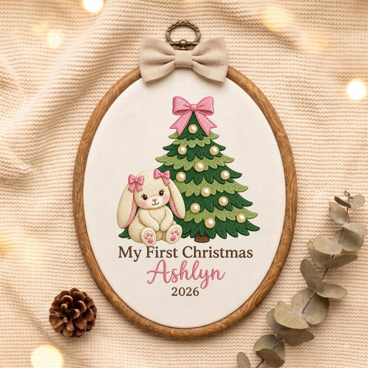 May include: An oval wooden embroidery hoop containing white fabric with a stitched design of a cream-colored bunny with pink bows, a green Christmas tree with pearl-like ornaments and a pink bow, and the text My First Christmas Ashlyn 2026.