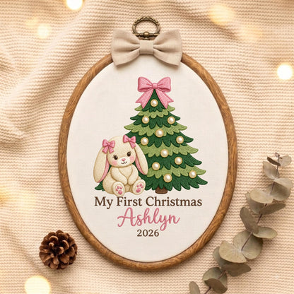 May include: An oval wooden embroidery hoop containing white fabric with a stitched design of a cream-colored bunny with pink bows, a green Christmas tree with pearl-like ornaments and a pink bow, and the text My First Christmas Ashlyn 2026.