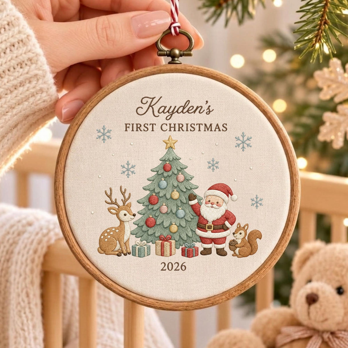 May include: A round wooden embroidery hoop ornament featuring a cream-colored fabric center with embroidered text reading Kayden's FIRST CHRISTMAS 2026. The design includes a decorated Christmas tree, Santa Claus, a deer, a squirrel, and snowflakes.