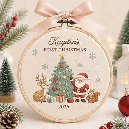 May include: A round wooden embroidery hoop featuring a cream fabric center with the embroidered text Kayden's FIRST CHRISTMAS and 2026. The design includes a decorated Christmas tree, Santa Claus, a deer, a squirrel, and snowflakes, topped with a pale pink ribbon.