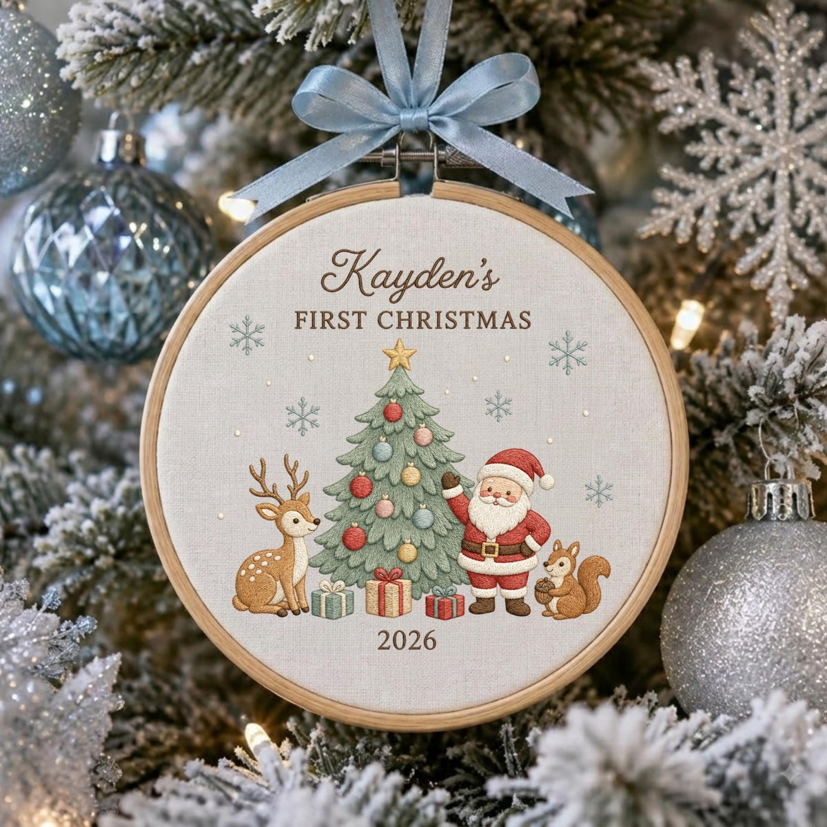 May include: A round wooden embroidery hoop ornament featuring a light fabric center with embroidered text Kayden's FIRST CHRISTMAS and 2026. The design includes a decorated Christmas tree, Santa Claus, a deer, a squirrel, and snowflakes.