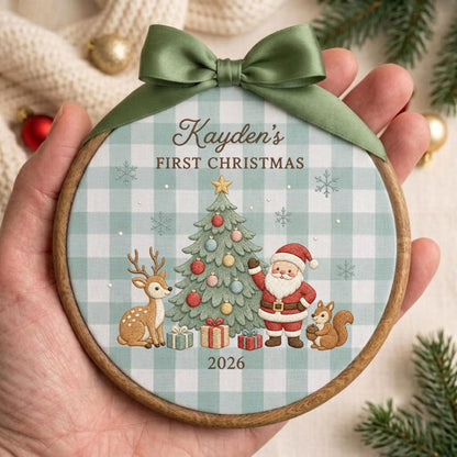 May include: A round wooden embroidery hoop ornament featuring a light blue and white buffalo check fabric background. The design includes a decorated Christmas tree, a deer, a squirrel, and Santa Claus with the text Kayden's FIRST CHRISTMAS 2026.