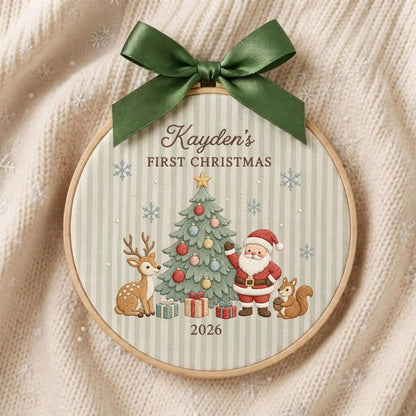 May include: A round wooden embroidery hoop ornament featuring a light green striped fabric background. The text reads Kayden's FIRST CHRISTMAS 2026. It depicts a decorated Christmas tree, Santa Claus, a deer, and a squirrel, topped with a dark green ribbon.